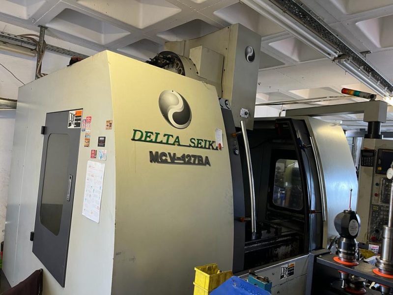 Endüstriyel bir atölyede kurulu durumda olan sarımsı beyaz renkli Delta Seiki MCV-127BA model ağır hizmet tipi döküm gövdeli CNC dik işleme merkezi takım tezgahının dıştan görünümü.